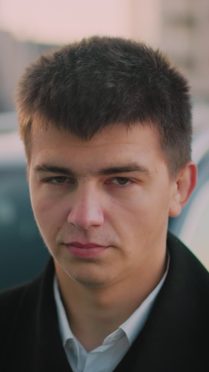 Man standing close to car with stern look, blurred background shows parked cars, distant building at dusk, with moving blurred figure in background