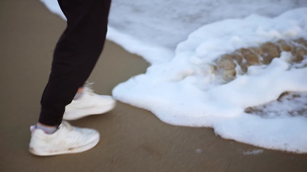 Man's legs in white sneakers standing on a sandy shore with waves coming at his feet