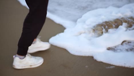 man's legs in white sneakers standing on a sandy shore with waves coming at his feet