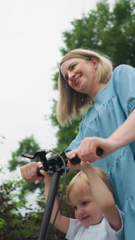 a mother and son ride a scooter together, both smiling warmly, close to a greenery background