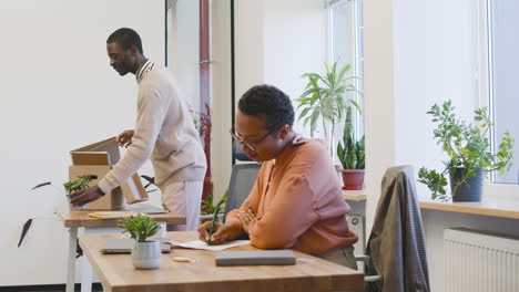 Woman-Sitting-At-A-Table-In-The-Office,-While-A-Young-Man-Takes-Out-His-Personal-Items-From-A-Box-And-Places-Them-On-The-Table