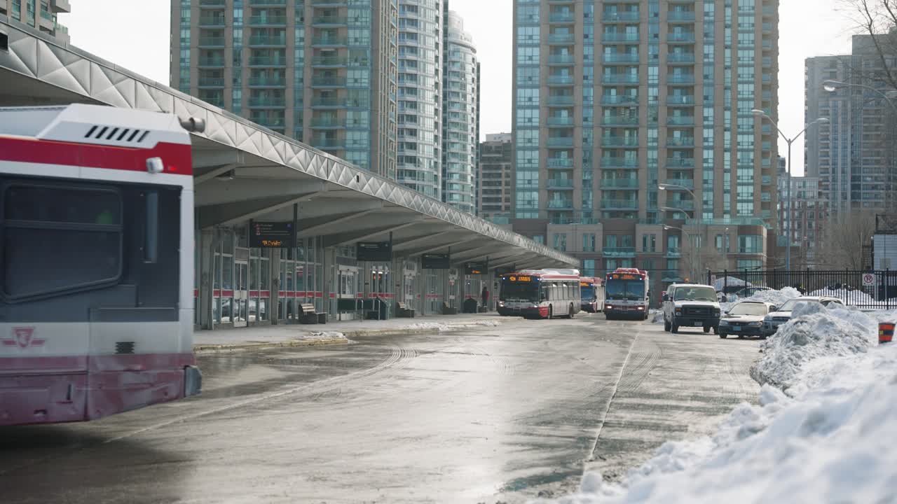Bus driving off from a bus station in winter, buildings in the background. Snow banks near by.