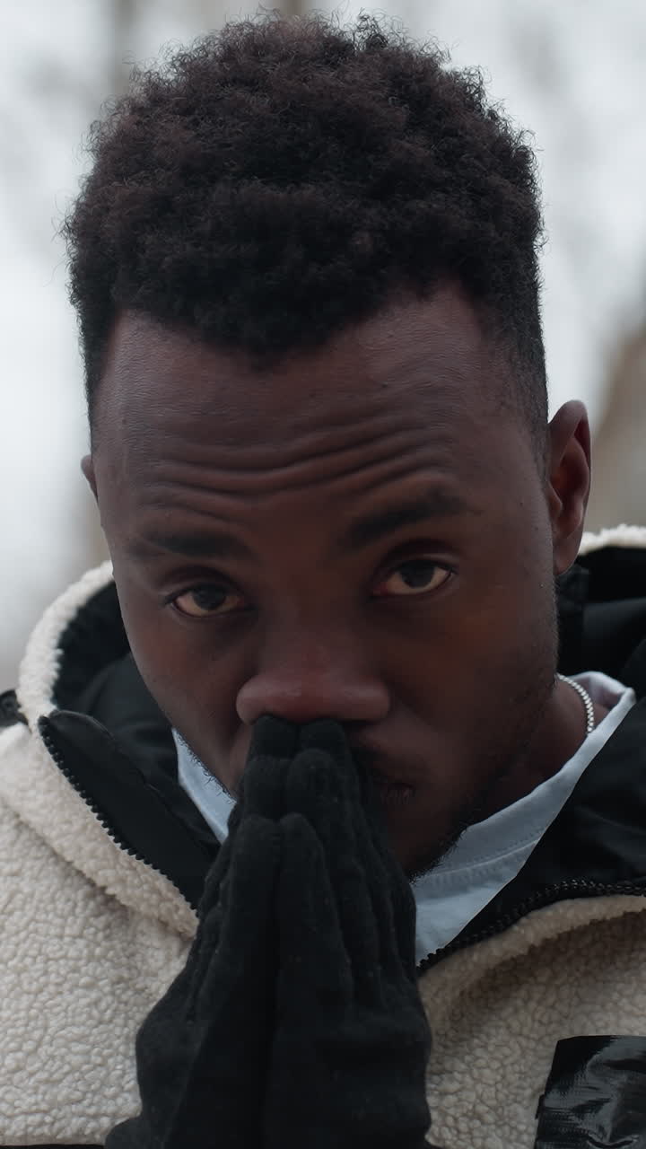 Man wearing black gloves and white winter jacket outdoors in cold weather, hands pressed together near face, thoughtful expression with blurred trees and structures in background