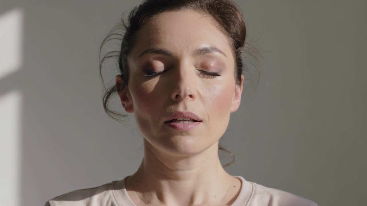 Mujer inhalando con camiseta de cuello redondo pálida en casa, ojos cerrados, labios entreabiertos, asintiendo con una sombra tenue