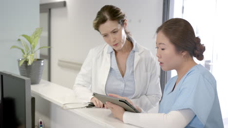 focused diverse female doctors using tablet and discussing at hospital reception desk, slow motion