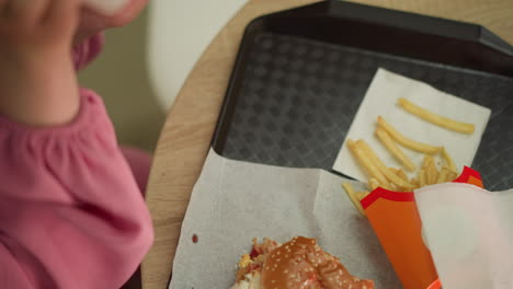 close-up of fries, burger, and coffee cup on a black tray on a brown wooden table, as a woman in a pink dress drops drink back onto the tray holds a half-eaten fry in her hand