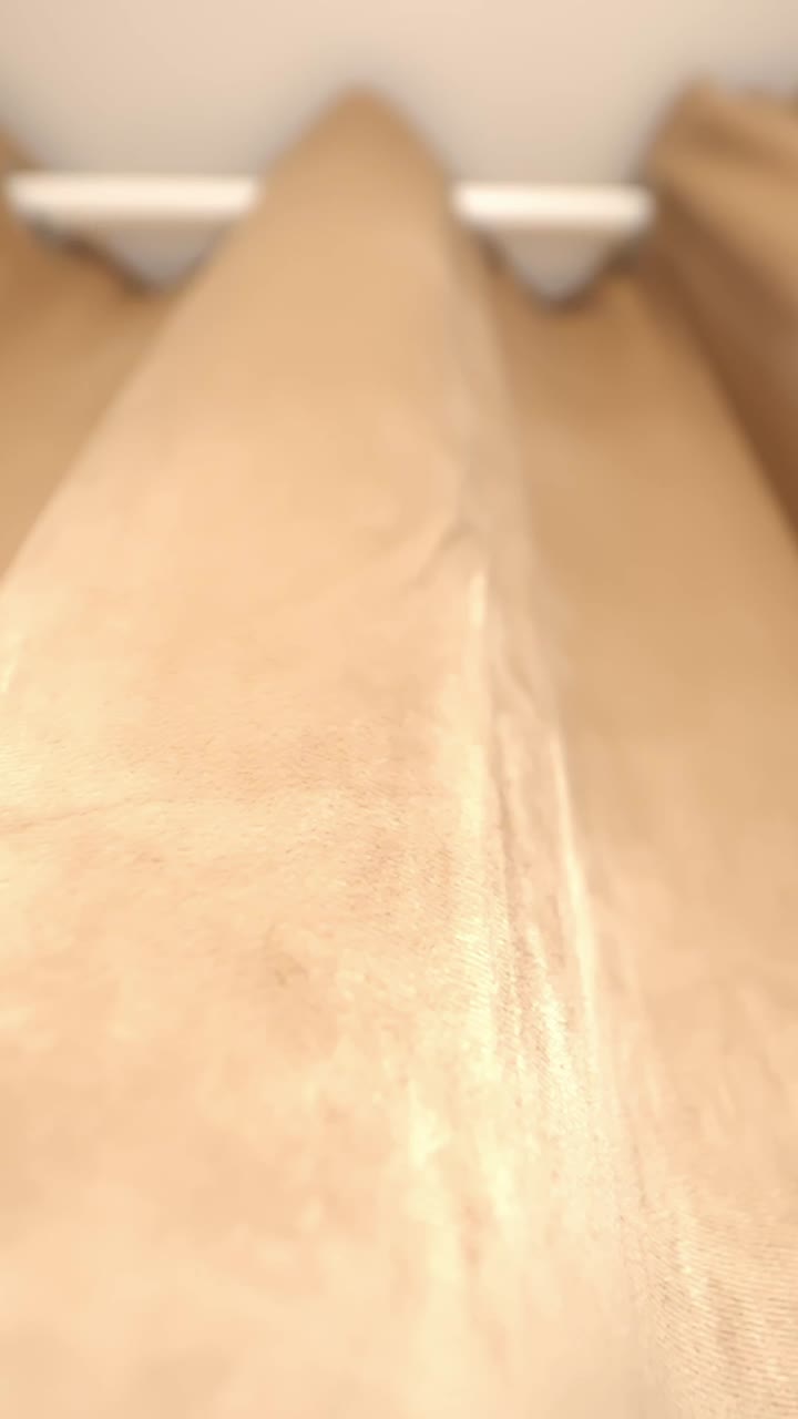 Ripresa verticale dal basso verso l'alto di tende beige con luce diurna calda che crea un'atmosfera interna morbida e minimale.