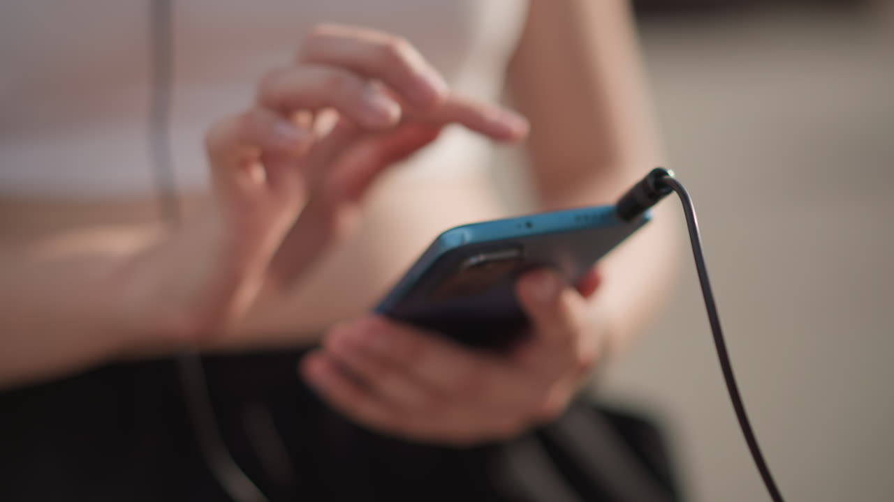 Female hands street holding phone plugged headphones, closeup of fingers tapping playlist app and scrolling tracks, wired cable visible against crop top and joggers, shallow depth of field