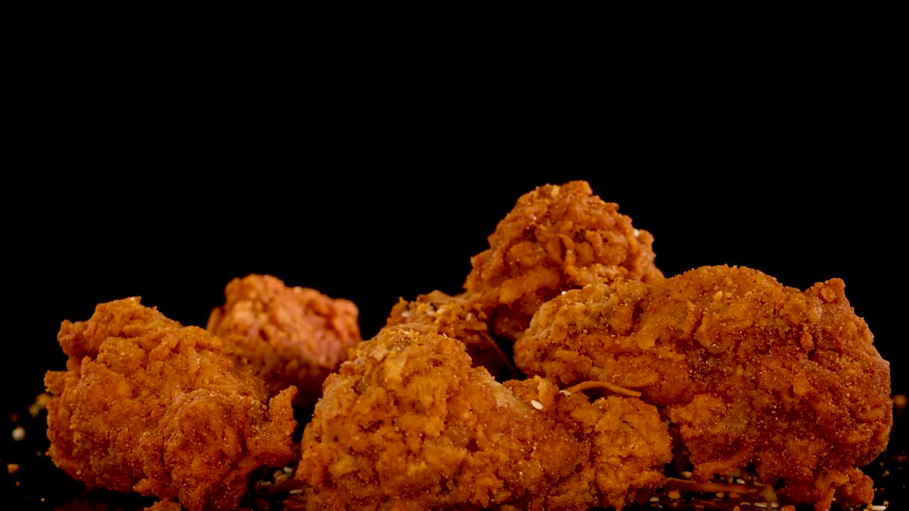 A hand reaches in to pick up a piece of spicy, crumbed fried chicken from a pile on a black background, shot in slow motion with dramatic lighting