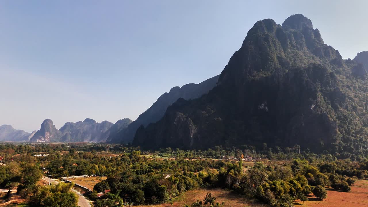 Mountainous terrain in Vang Vieng, Laos featuring dramatic limestone cliffs, a forested valley, distant ridges, and a small settlement under clear blue skies during daylight
