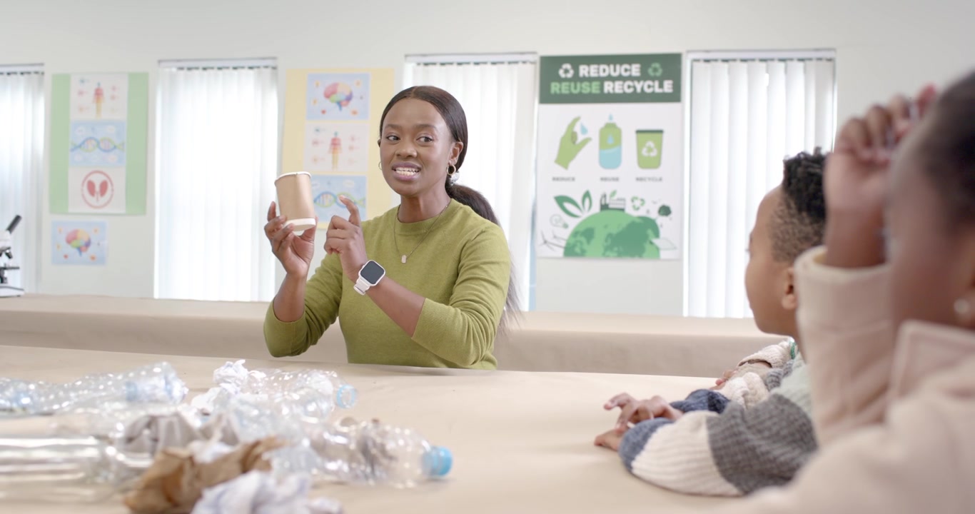 Teacher leading kids reaching into pile and lifting paper cup at classroom table teaching recycling