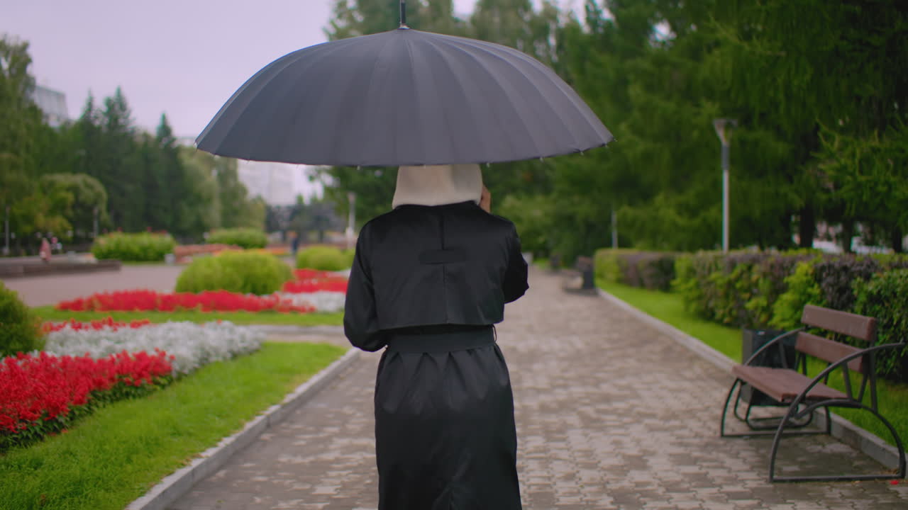 Woman in black coat with hood walks under umbrella on rainy park path, seen from behind, surrounded by red and white flowers, green trees and cloudy sky creating calm urban rainy day mood