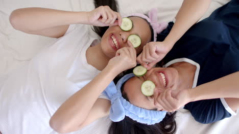 two biracial teenage girl friends lying down at home, holding cucumber slices over their eyes