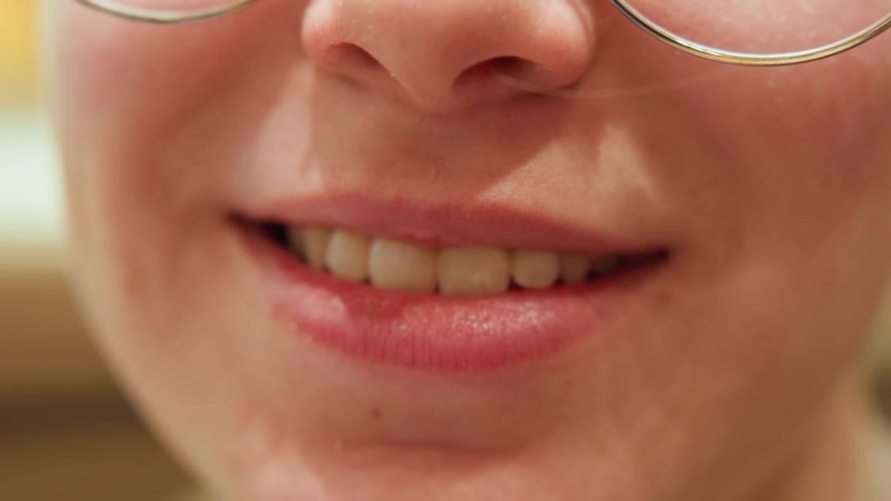 Primo piano di un narratore caucasico che parla e ride, le labbra si muovono da un bacio a labbra serrate a un discorso aperto e a un sorriso. Montatura degli occhiali visibile, articolazione dinamica della bocca. Scena spontanea e calda che suggerisce una prova.