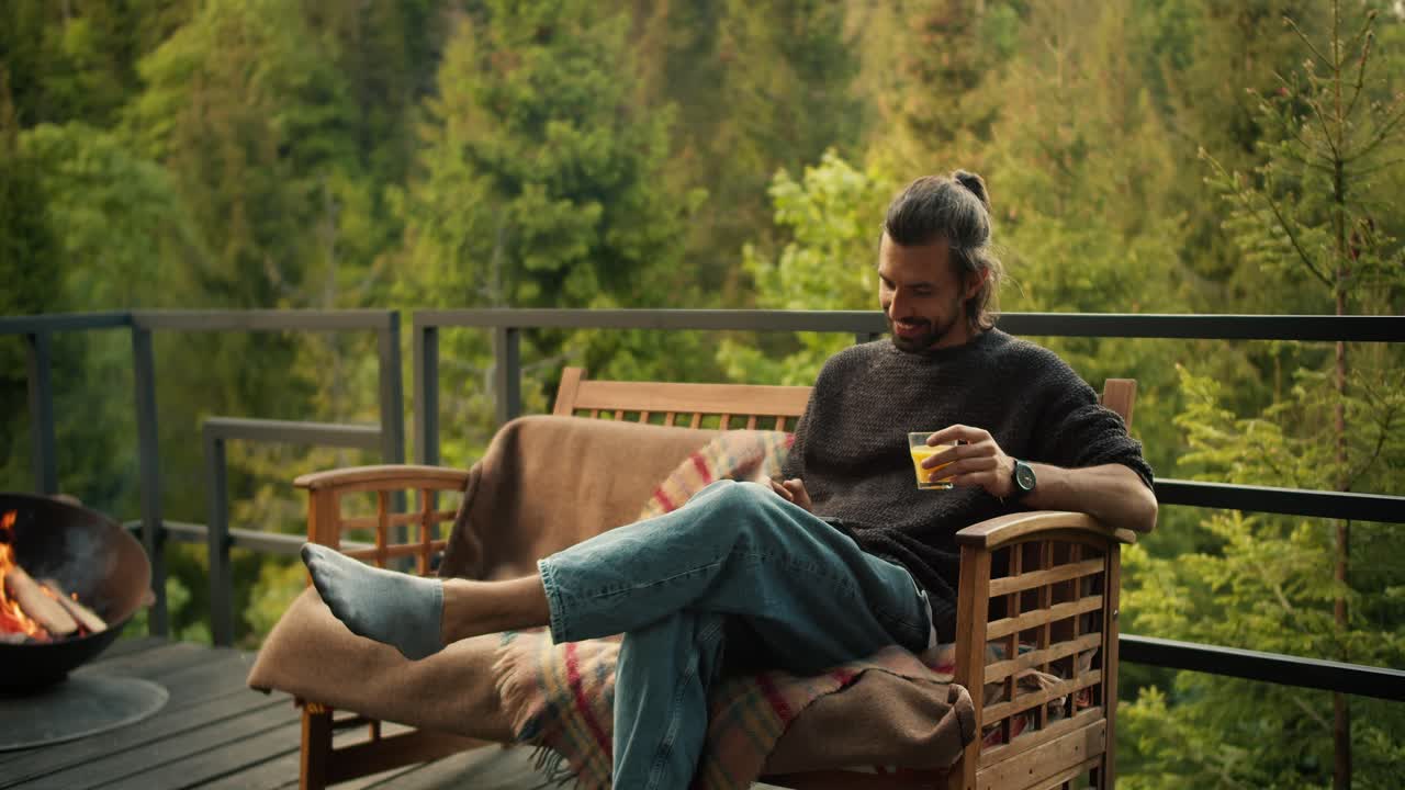 A man with a yellow drink in his hand sits on a sofa and works on a tablet on the balcony of a country house overlooking the mountains and coniferous forest