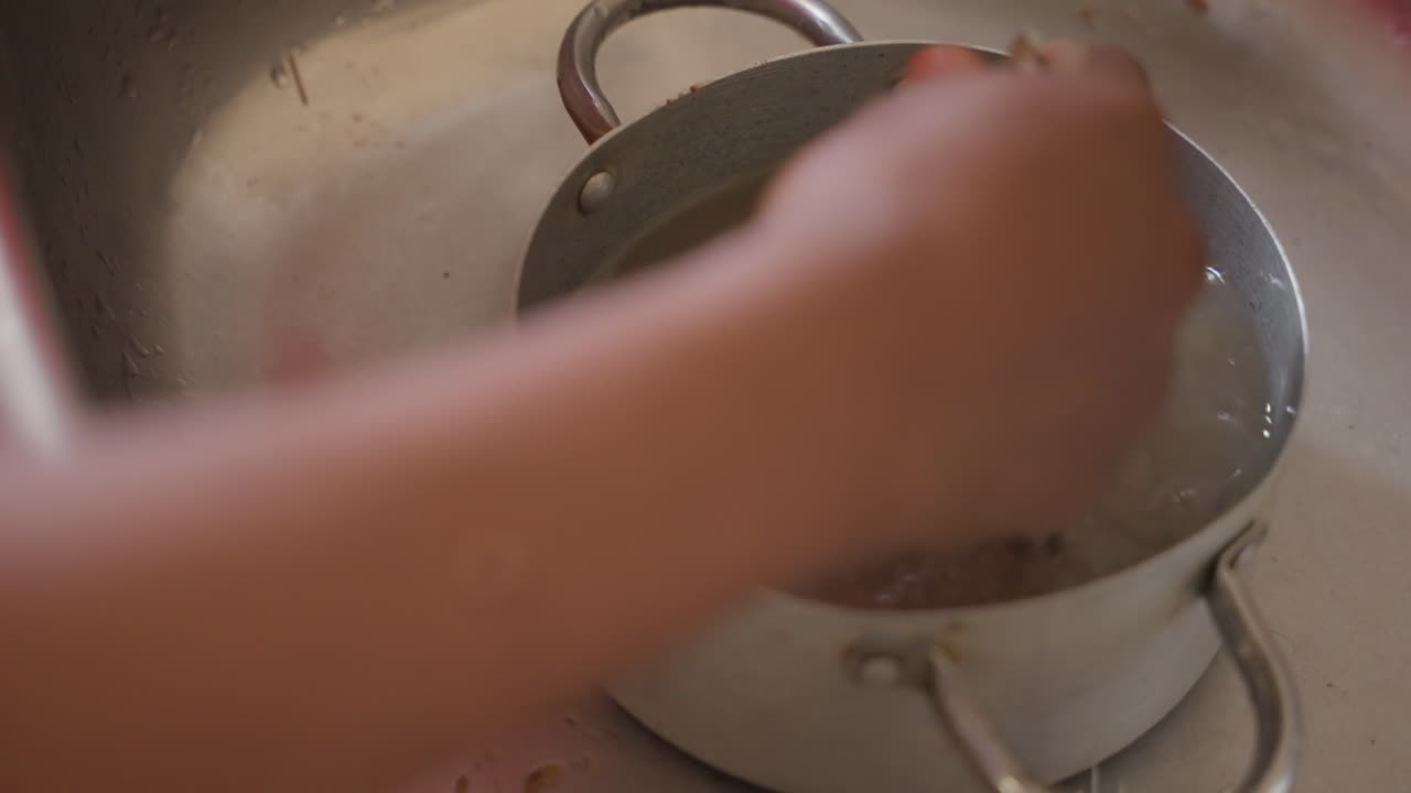 Kitchen sink cleaning. Methodical hand wash of stained pots. Washing greasy pots in stainless steel sink carefully. Meticulous scrubbing of muddy pots with circular motions in kitchen sink