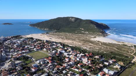 Aerial-drone-view-of-tropical-beach-with-vegetation-mountains,-green-fields,-blue-ocean-and-urban-road-network,-urban-streets-at-Santinho-beach-beach-houses-and-urbanism