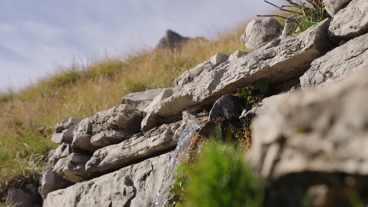 Clear water flows down a small waterfall emerging from rocks on a grassy hill