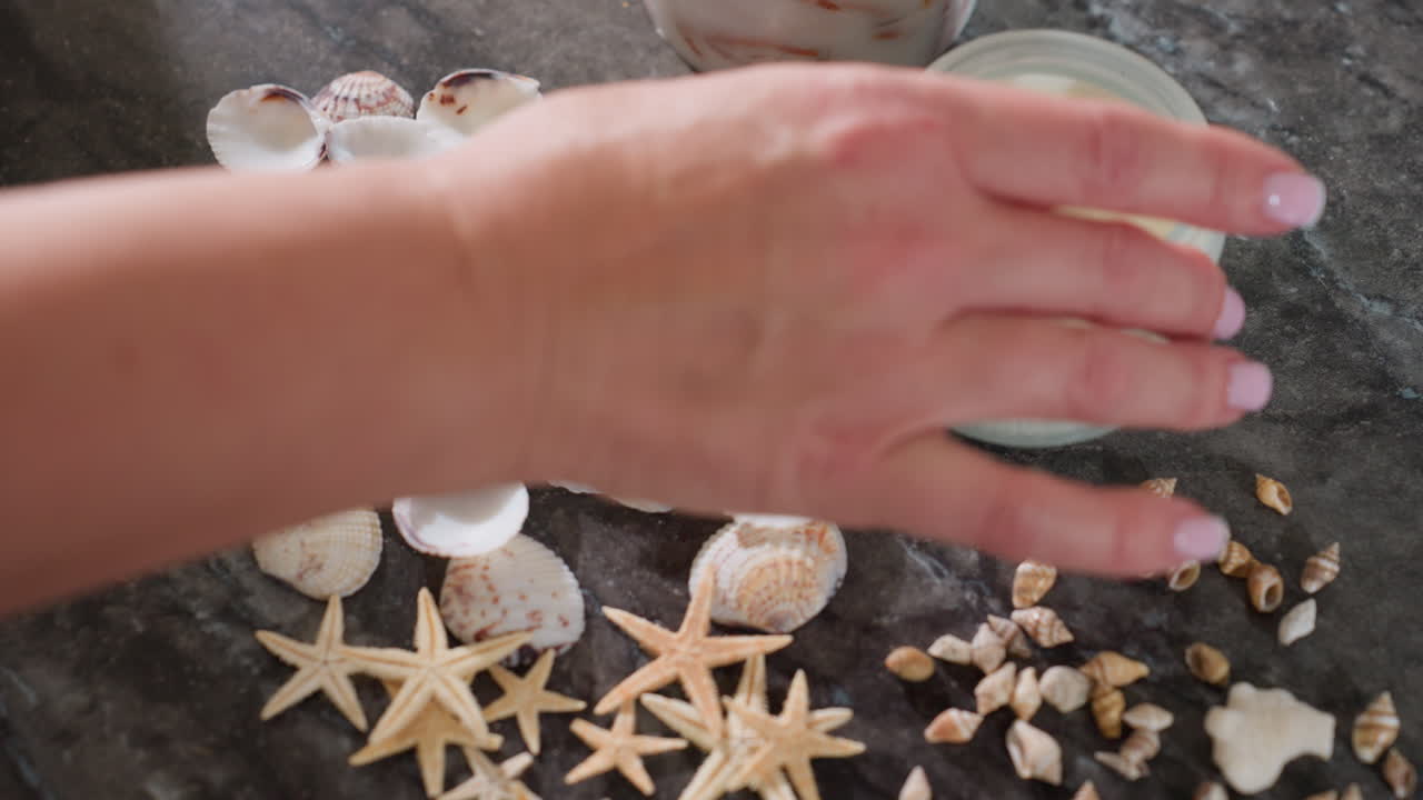 Aerial view of lady adjusting starfish seashells and snail shells around white candle jar on marble tabletop creating decorative beach inspired arrangement