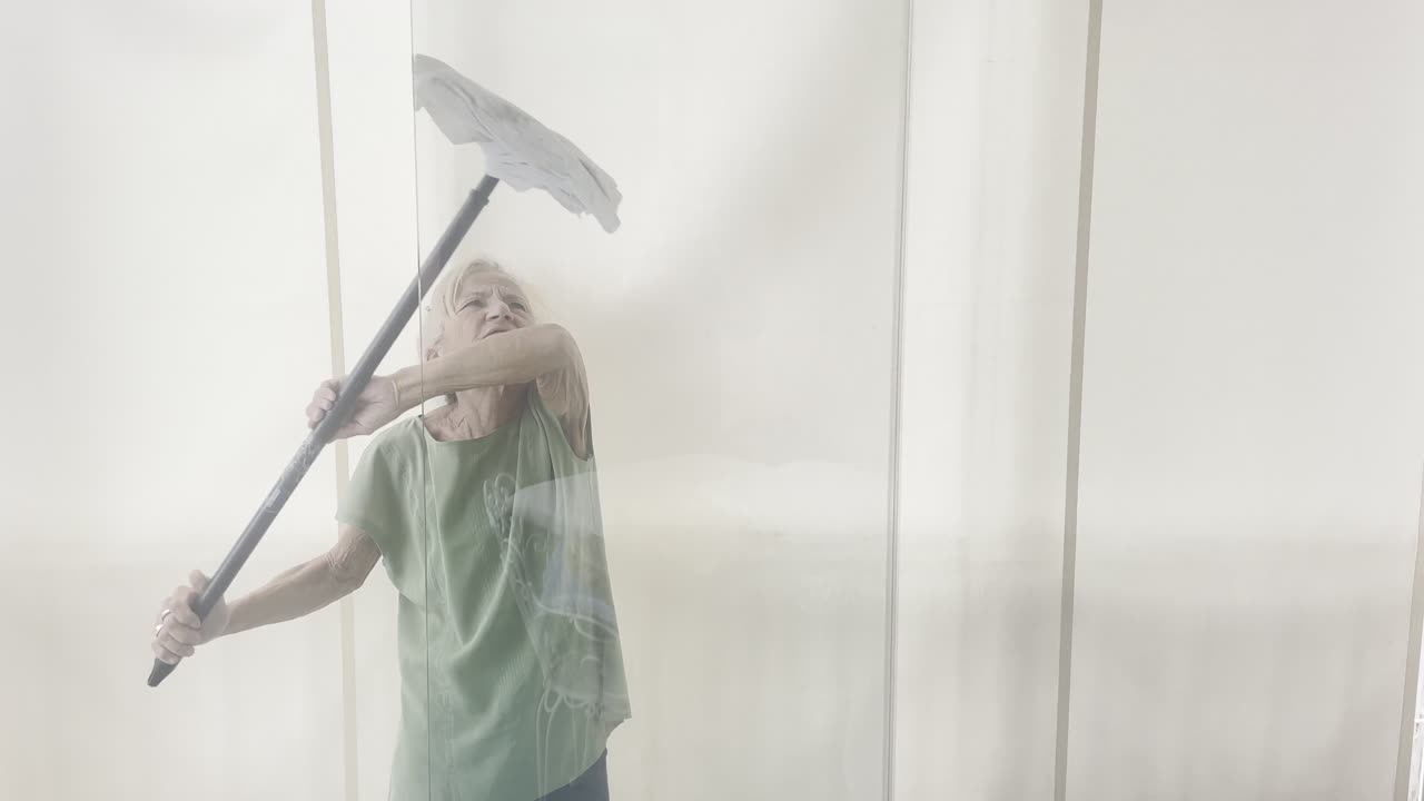 Oudere vrouw gebruikt een trekker met lange steel om een hoog glazen raam schoon te maken. Door de weerspiegeling is te zien hoe ze met inspanning en focus werkt in een lichte huiselijke omgeving
