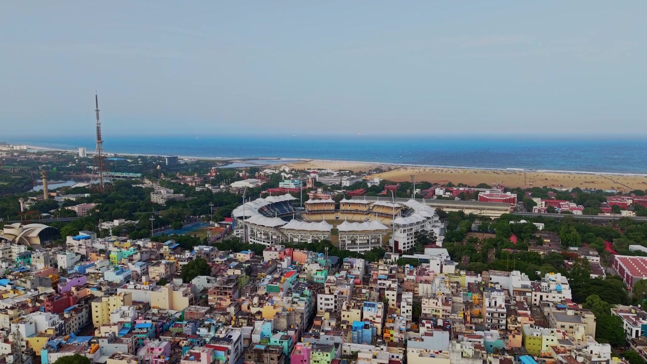 Ripresa aerea con drone che mostra uno stadio di cricket circondato da edifici densi e vegetazione, con Marina Beach e la costa del Golfo del Bengala a Chennai, Tamil Nadu, India