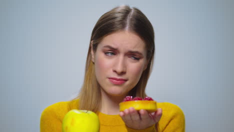 portrait of thoughtful woman selecting between apple or cake at camera.