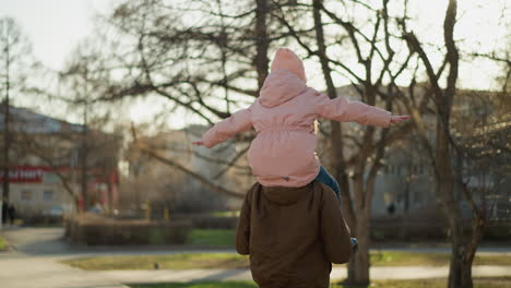 father walks through park with little girl on his shoulders, her arms spread wide in joy, both in pink cap and jacket