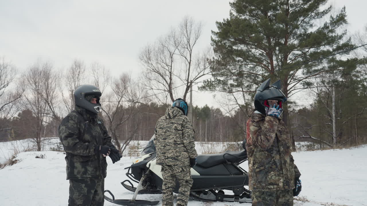 White riders by snowmobile and ATV in snowy forest, three camouflaged riders in helmets converse beside parked snowmobile and distant ATV, guide assigning trail roles, visible breath in cold air, pine