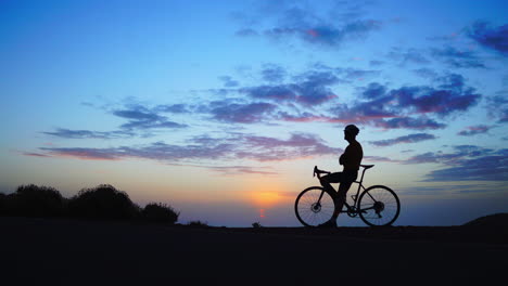 El-Hombre-Está-Parado-Junto-A-Una-Bicicleta,-Enmarcado-Contra-La-Puesta-De-Sol-De-Fondo.-El-Momento-Se-Captura-En-Una-Secuencia-De-Lapso-De-Tiempo,-Utilizando-Una-Vista-De-Gran-Angular