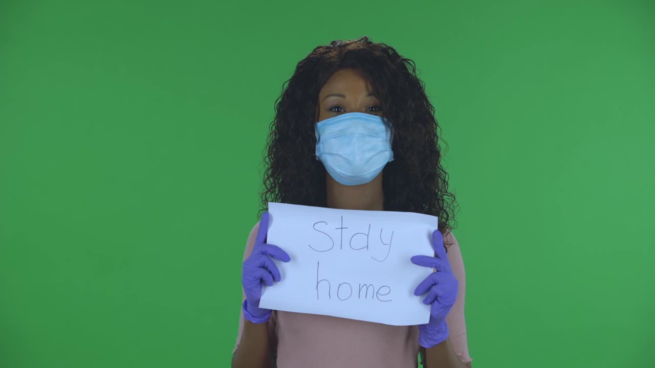 Portrait of beautiful african american young woman in medical mask is looking at camera and holding a poster with the words stay home. Burning brunette with wavy hair in jeans and a beige blouse on a green screen in the studio. Health Protection Corona