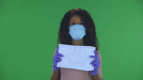 portrait of beautiful african american young woman in medical mask is looking at camera and holding a poster with the words stay home. burning brunette with wavy hair in jeans and a beige blouse on a green screen in the studio. health protection corona