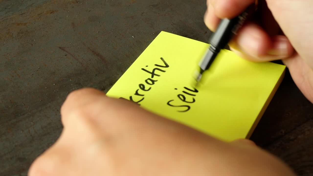Hand writing "Kreativ Sein" (Creative Being) on a yellow sticky note