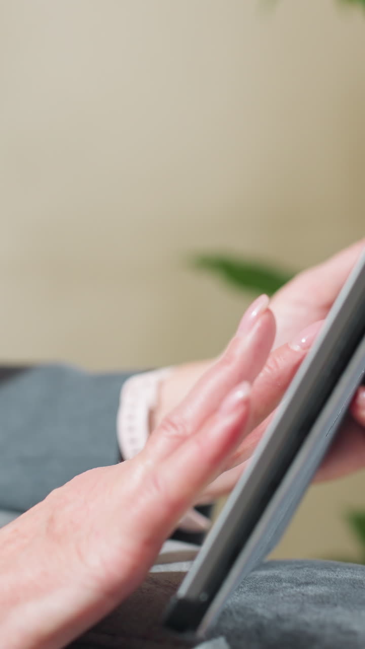 Businesswoman in suit sitting comfortably while using tablet indoors near green plant, focusing on screen for digital task, online communication, or remote work in relaxed professional environment