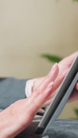 businesswoman in suit sitting comfortably while using tablet indoors near green plant, focusing on screen for digital task, online communication, or remote work in relaxed professional environment