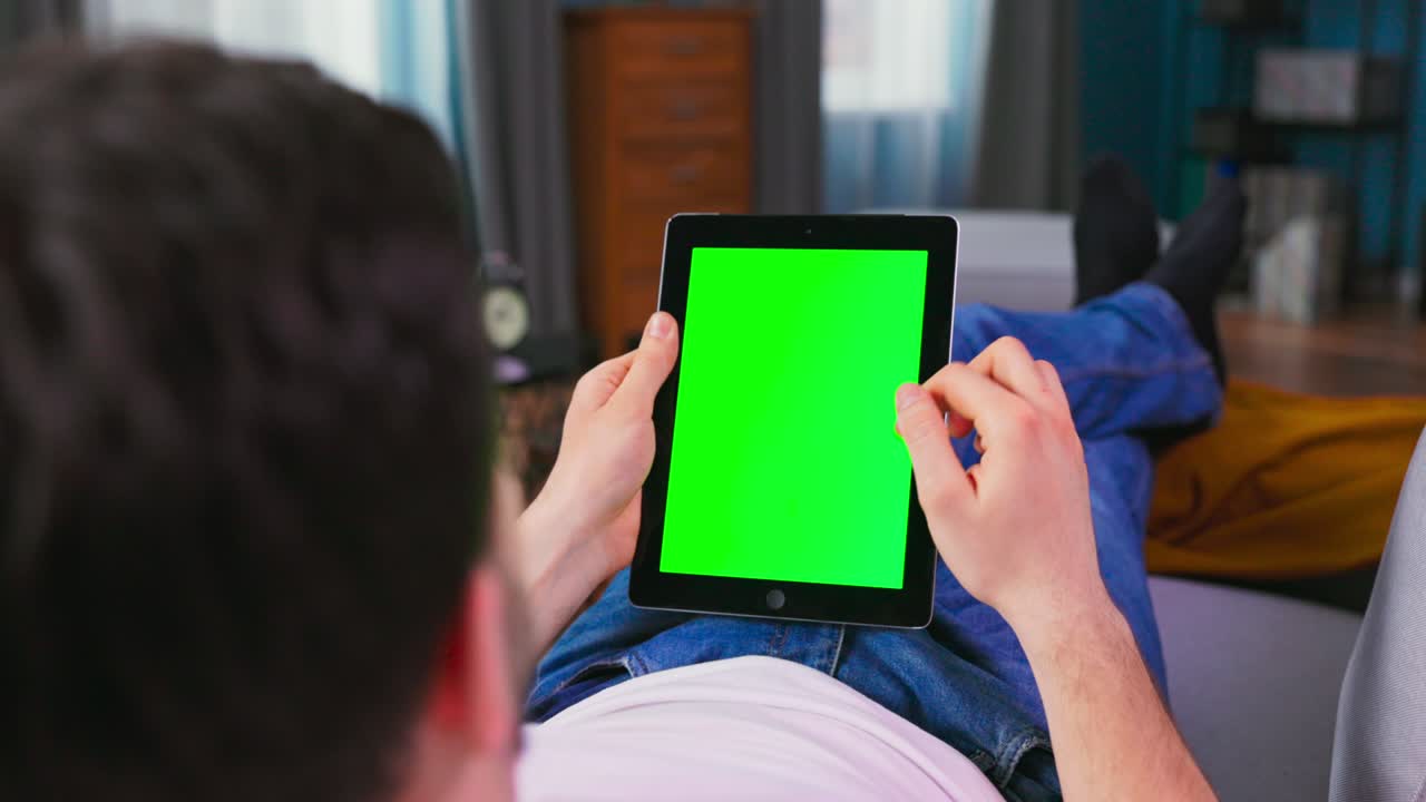 Student boy Resting on a Couch Using with Green Mock-up Screen Tablet Computerin Vertical Portrait Mode. Man Using Gestures with Touchscreen Device, Browsing Internet, Watching Content