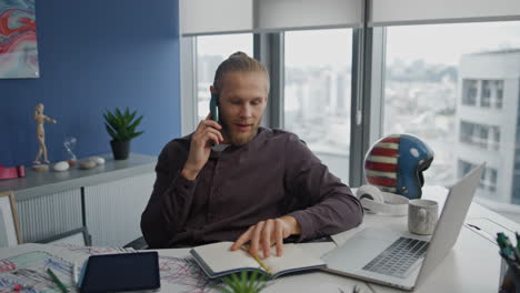 Hombre-Sonriente-Charlando-Con-Un-Teléfono-Inteligente-En-El-Primer-Plano-De-La-Oficina.-Cuaderno-De-Observación-De-Startups