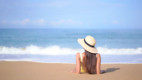 Junge-Frau-Liegt-Am-Sandstrand-Am-Meer-Und-Stützt-Sich-Auf-Ellbogen-Mit-Sonnenhut-Und-Gelbem-Badeanzug---Rückansicht