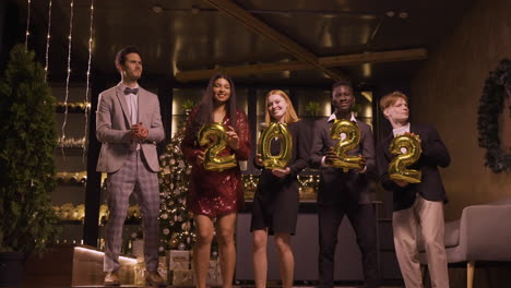 group of friends wearing elegant clothes holding balloons with the numbers 2022 while dancing and hugging each other in new year's party