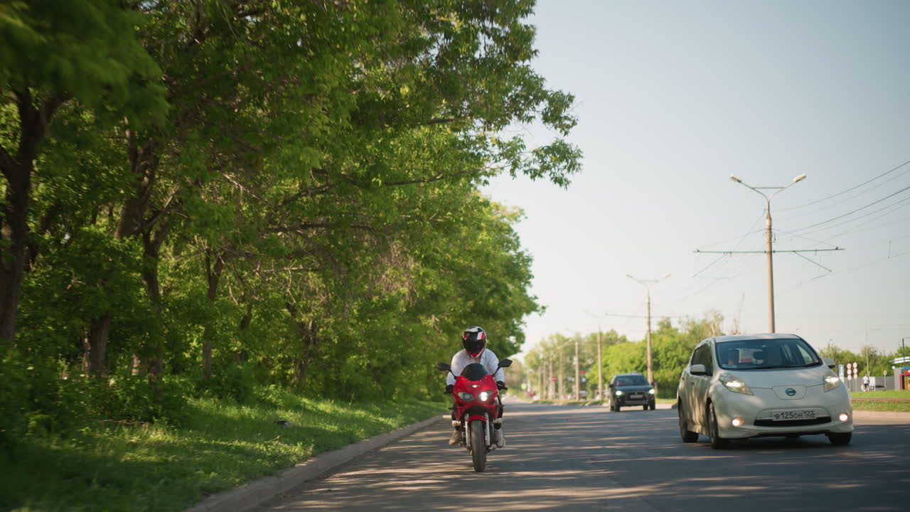 한 여성 오토바이 운전자가 한 개의 헤드라이트를 켜고 빨간색 파워바이크를 타고 도시 환경에서 울창한 초록색 나무 근처를 지나가고 자동차가 그녀를 가게 지나가며 다른 차량이 다른 차선에서 볼 수 있습니다.