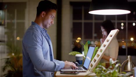 biracial casual businessman working on laptop at standing desk at night in office, in slow motion