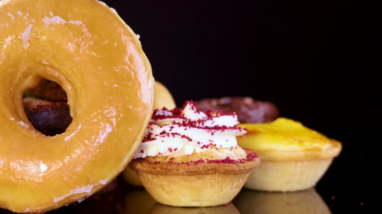 A variety of desserts including glazed donuts, cupcakes, brownies, tarts, and strawberries rotate under bright studio lighting against a dark, reflective backdrop