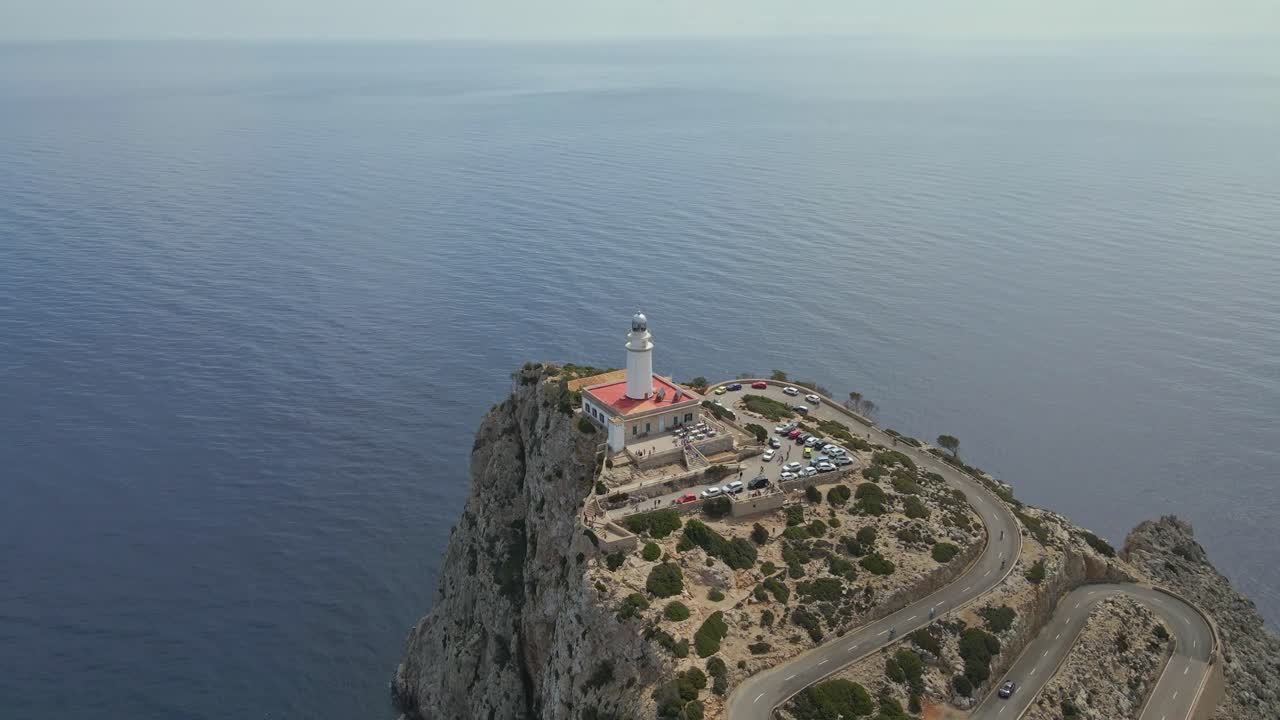 An aerial video of a lighthouse perched on a rugged cliffside overlooking the calm blue sea. The winding road and parked cars suggest it is a popular tourist destination with stunning coastal vistas.