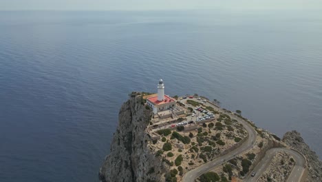 An aerial video of a lighthouse perched on a rugged cliffside overlooking the calm blue sea. The winding road and parked cars suggest it is a popular tourist destination with stunning coastal vistas.