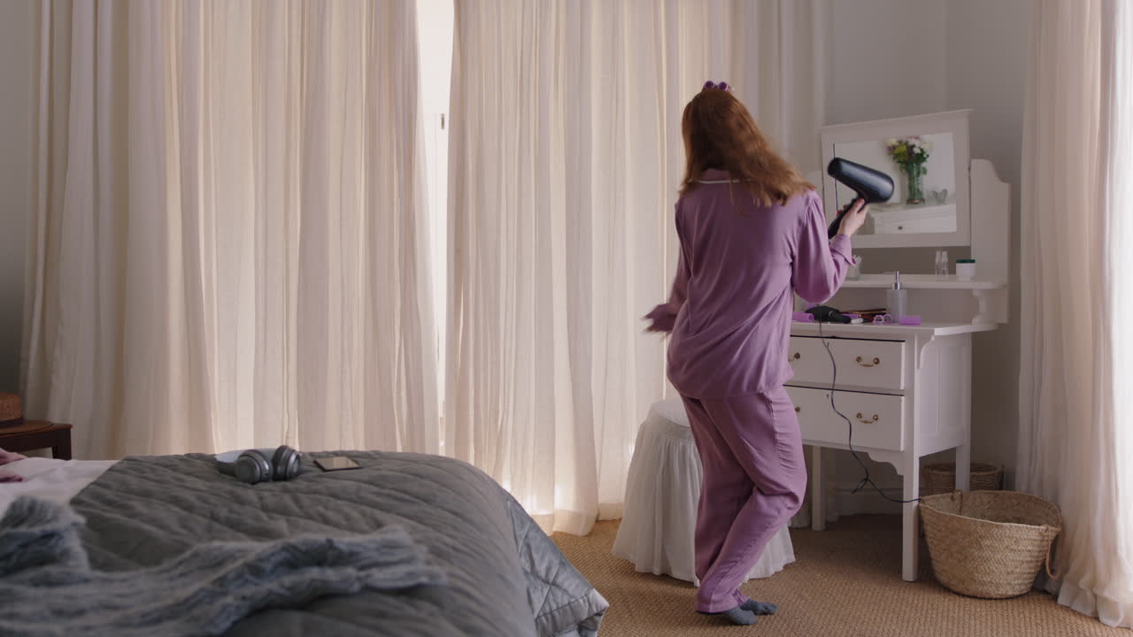 재미있는 여자가 침실에서 춤을 추고 머리카락 건조기와 함께 카라오케를 부르고 주말 아침을 즐기고 즐겁게 즐기고 집에서 피자마를 입고 무관심한 라이프 스타일을 축하합니다.