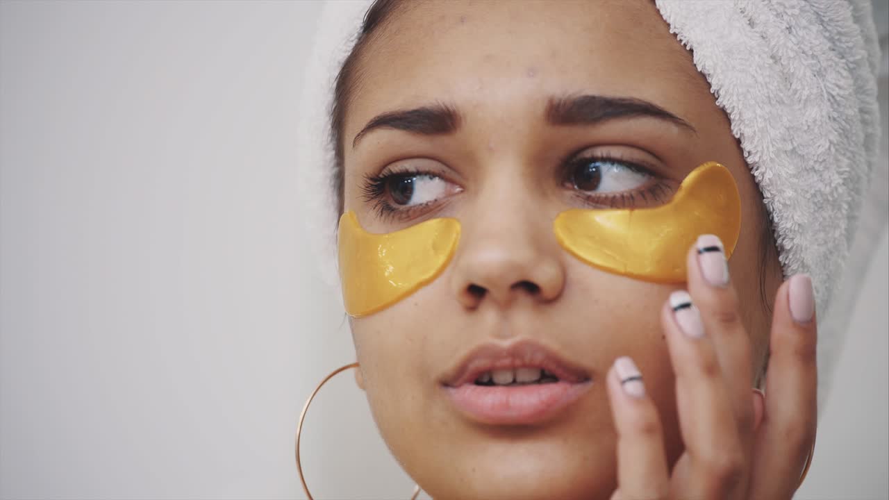 Closeup portrait of beautiful woman after bath with towel on head puts patches under the eyes from wrinkles and dark circles. Attractive female in bathrobe makes beauty treatments on the face at home.