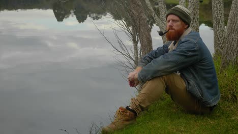 a mid shot of a bearded ginger mountain man smoking a tobacco pipe while sitting by a calm lake