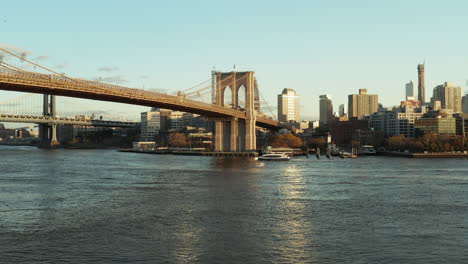 Low-flight-above-water,-cruise-ship-floating-on-surface-under-Brooklyn-Bridge.-High-rise-buildings-in-background.-Brooklyn,-New-York-City,-USA