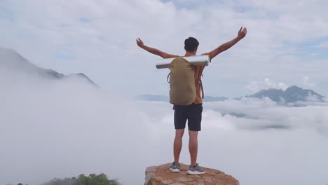 asian hiker male reaching up top of foggy mountain standing on the rock and raising his hands