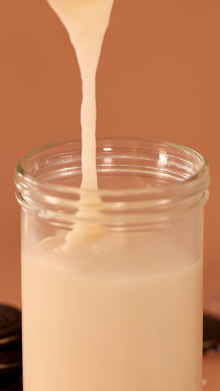 Primo piano al rallentatore di un denso milkshake alla vaniglia che si versa in un barattolo di vetro su uno sfondo marrone caldo.