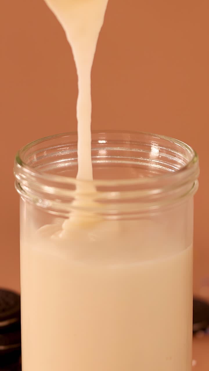 Primo piano al rallentatore di un denso milkshake alla vaniglia che si versa in un barattolo di vetro su uno sfondo marrone caldo.